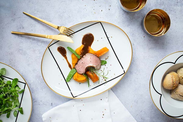 Ein elegant angerichteter Teller mit gebratenem Fleisch, Kürbispüree und Zuckerschoten, flankiert von goldenem Besteck und passendem Geschirr auf einer grauen Steinfläche.
