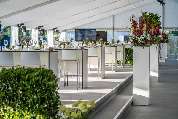 Festlich eingedecktes Eventzelt mit modernen weißen Stehtischen, Barhockern und hoher Blumendekoration.