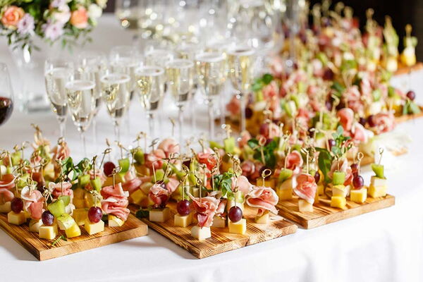 Ein Buffet mit reichlich angerichteten Fingerfood-Spießen auf Holzbrennbrettern und gefüllten Sektgläsern im Hintergrund.
